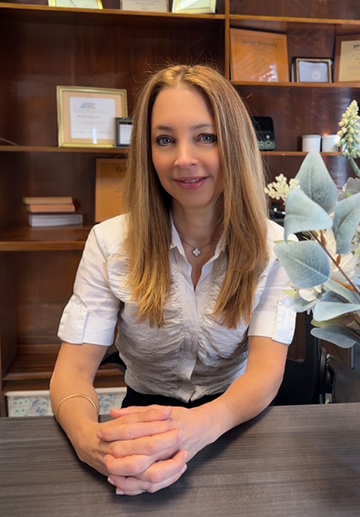 A woman with long, straight hair sits at a desk, hands folded, wearing a white blouse. Bookshelves with certificates and plants are in the background.
